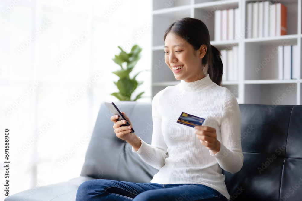 Happy young Asian woman using smartphone and credit card for online shopping on sofa at home. Happy young Asian woman using smartphone and credit card for online shopping on sofa at home.