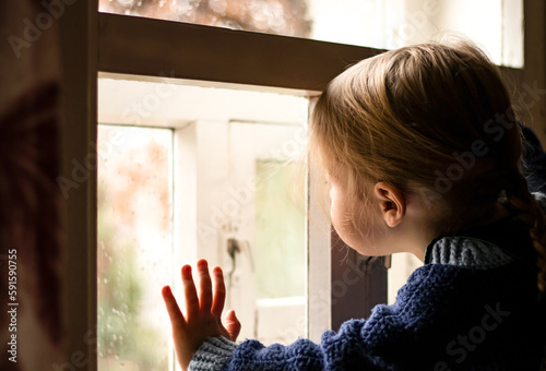 little child girl looking through window in raindrops at home holding hand on glass, it's raining outside, rainy day, cloudy weather photo