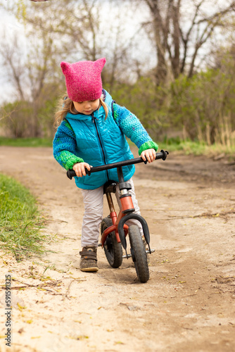 Fototapeta Naklejka Na Ścianę i Meble -  little child girl in blue jacket and pink hat having fun riding on bicycle on the road outdoors