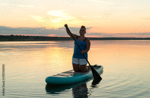 A woman on a sapboard on her knees with an oar at sunset against the sky floats in the water of the lake. © finist_4