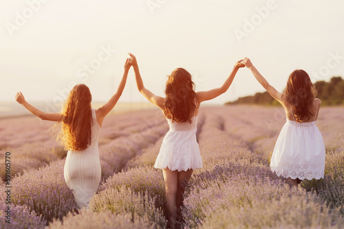 Young girls in a white dresses walking joyfully through a lavender field and holding hands up under the rays of the setting sun.