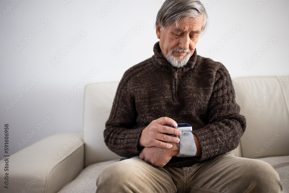 Senior man using medical device to measure blood pressure - elderly man suffering from high blood pressure sitting at home on sofa takes care of his health Senior man using medical device to measure blood pressure - elderly man suffering from high blood pressure sitting at home on sofa takes care of his health