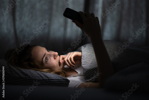 Young woman in bed holding a phone, tired and exhausted, blue light straining her eyes, messing up her circadian rhytm