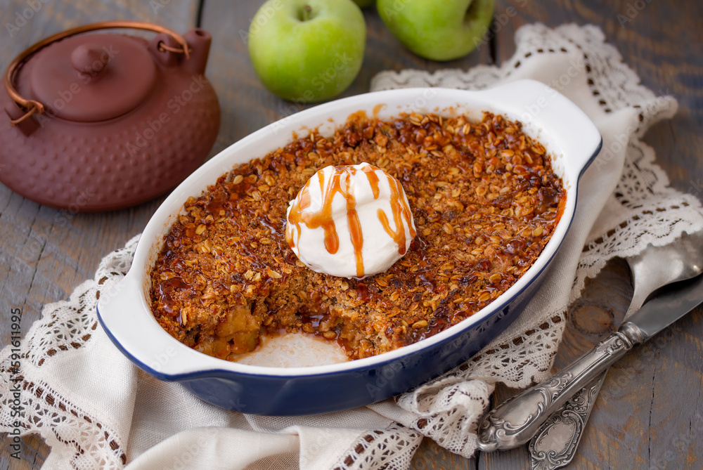 Homemade Apple Crisp or Apple Crumble with toasted crunchy streusel topped with vanilla ice cream and caramel sauce served in baking dish with kettle and fresh green apples on background. Horizontal. Homemade Apple Crisp or Apple Crumble with toasted crunchy streusel topped with vanilla ice cream and caramel sauce served in baking dish with kettle and fresh green apples on background. Horizontal.