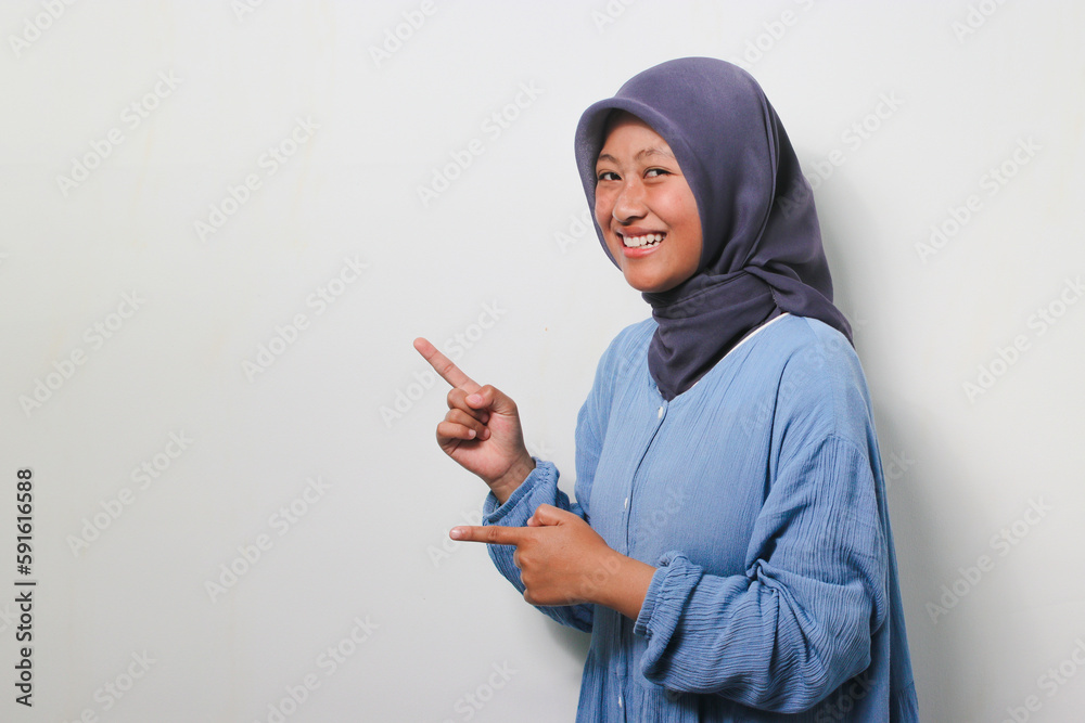 Excited young Asian girl in hijab pointing finger left on white background. Excited young Asian girl in hijab pointing finger left on white background.