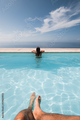 girl from behind looking at the sea inside an infinity pool with some man s legs in the foreground on a sunny day