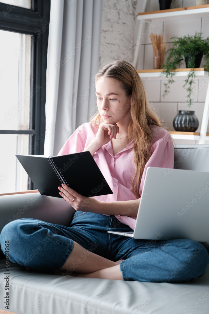 Thoughtful caucasian young woman in casual clothes is sitting cross-legged at home on the sofa with a laptop, looking at a planner, thinking, reading a text Thoughtful caucasian young woman in casual clothes is sitting cross-legged at home on the sofa with a laptop, looking at a planner, thinking, reading a text