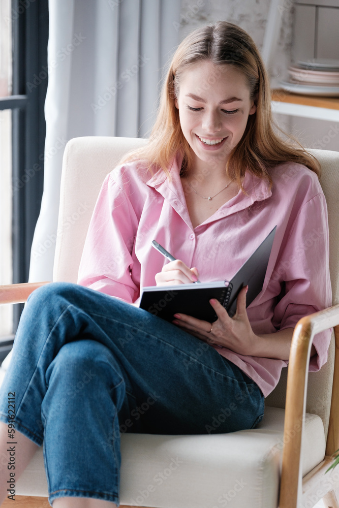 Laughing caucasian young woman in casual clothes is sitting at home in a chair with her legs crossed, making notes in a planer, writing a story in a book Laughing caucasian young woman in casual clothes is sitting at home in a chair with her legs crossed, making notes in a planer, writing a story in a book