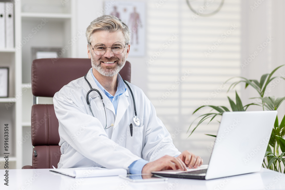 Portrait of a mature adult gray-haired doctor inside a medical office, the doctor is smiling and looking at the camera, the man is sitting at a table with a laptop and in a white medical coat. Portrait of a mature adult gray-haired doctor inside a medical office, the doctor is smiling and looking at the camera, the man is sitting at a table with a laptop and in a white medical coat.
