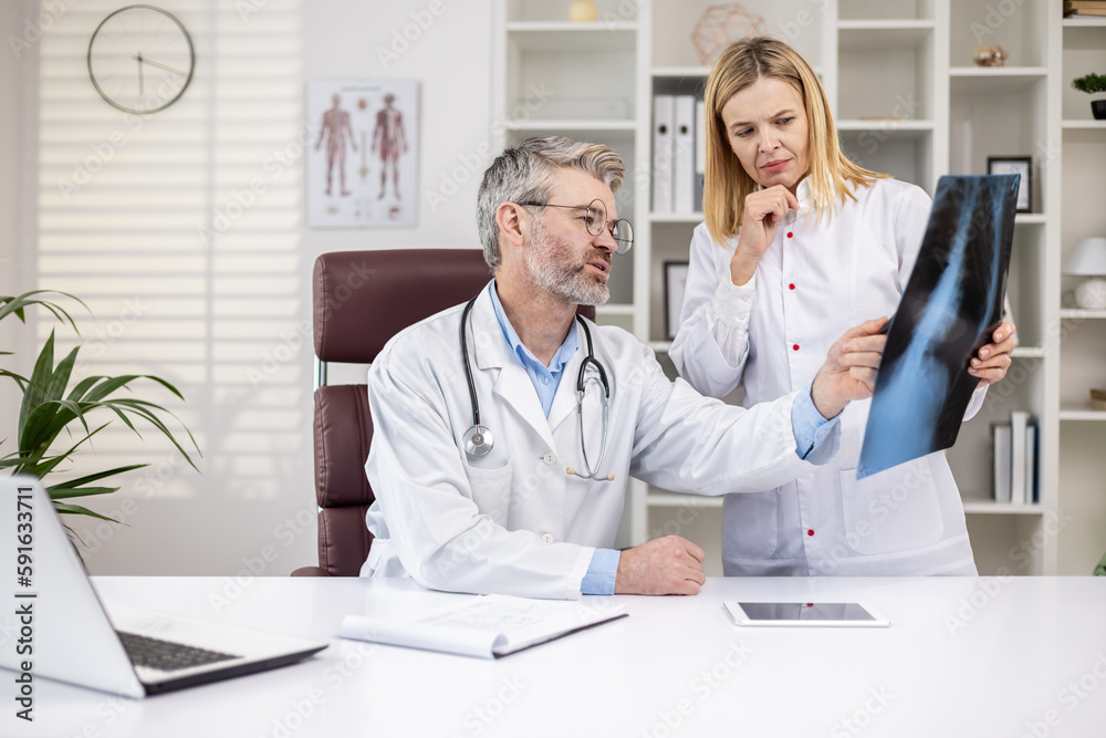 Serious mature doctors thinking and discussing patient's x-ray, man and woman in medical white coats working inside clinic office, working consult. Serious mature doctors thinking and discussing patient's x-ray, man and woman in medical white coats working inside clinic office, working consult.