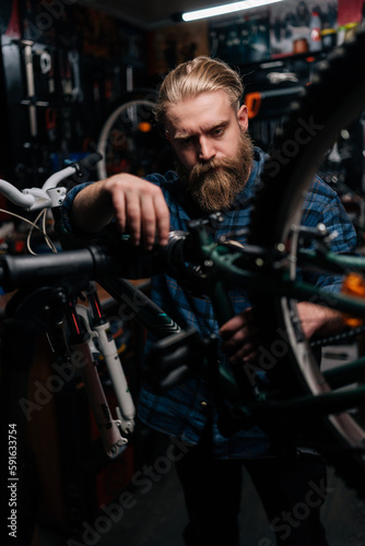 Vertical portrait of bearded brutal repairman repairing and fixing mountain bicycle standing on bike rack working in bike repair shop with dark interior. Concept of maintenance of bicycle transport.