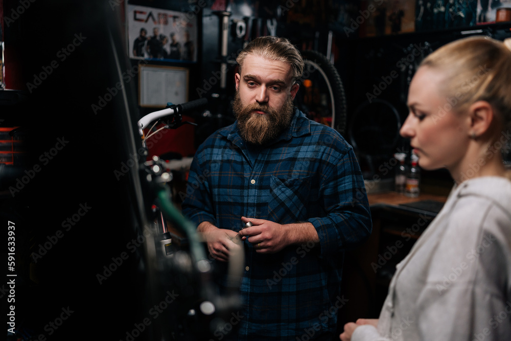 Side view of bearded cycling repairman having conversation with blonde female client, talking about problem of bicycle detected during diagnostics in repair shop with dark interior. Side view of bearded cycling repairman having conversation with blonde female client, talking about problem of bicycle detected during diagnostics in repair shop with dark interior.