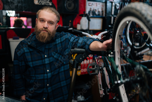 Portrait of handsome bearded cycling repairman standing by bicycle in repair bike workshop with dark interior, looking at camera. Concept of professional repair and maintenance of bicycle transport.
