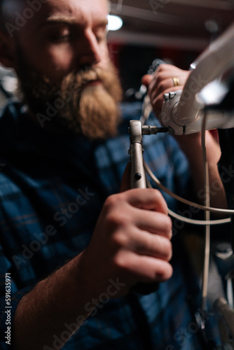 Closeup vertical shot of concentrated bearded repairman fixing bicycle handlebar with special tools working in bike repair shop with dark interior. Concept of professional maintenance of bike.