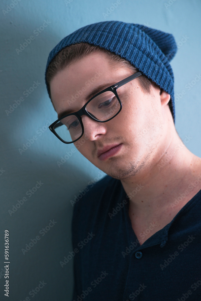 Close-up Portrait of Young Man wearing Woolen Hat and Horn-rimmed Eyeglasses, Looking Downward, Studio Shot on Blue Background Close-up Portrait of Young Man wearing Woolen Hat and Horn-rimmed Eyeglasses, Looking Downward, Studio Shot on Blue Background
