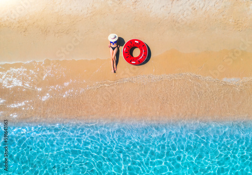 Aerial view of a lying woman in hat with red swim ring on sandy beach and blue sea at sunset in summer. Tropical landscape with girl, clear water, waves. Top view. Vacation. Sardinia island, Italy	