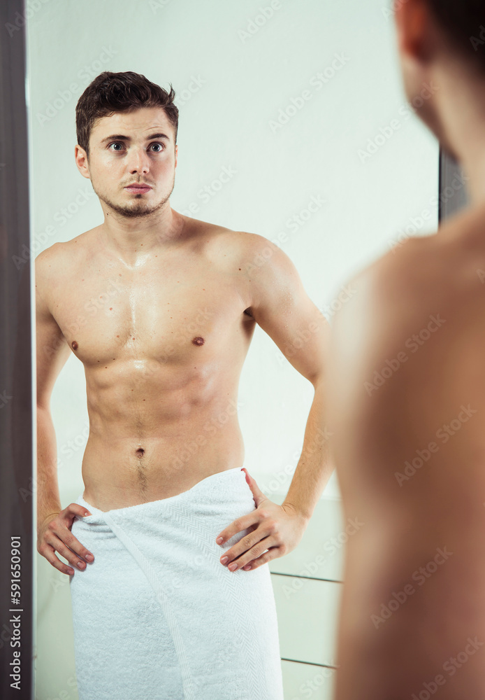 Portrait of young man with towel wrapped around waist, looking in bathroom mirror, studio shot Portrait of young man with towel wrapped around waist, looking in bathroom mirror, studio shot