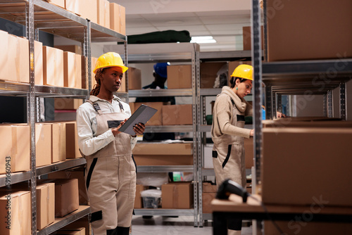 Warehouse manager supervising freight inventory and checking audit application. African american woman standing in storehouse near cardboard boxes shelf and using digital tablet