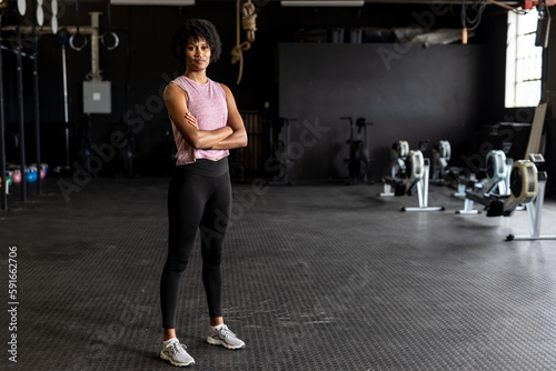 Full length portrait of biracial confident young woman with arms crossed standing in gym