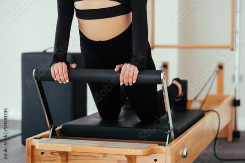 Unrecognizable woman wearing black sportswear practicing pilates exercises on reformer machine in studio.