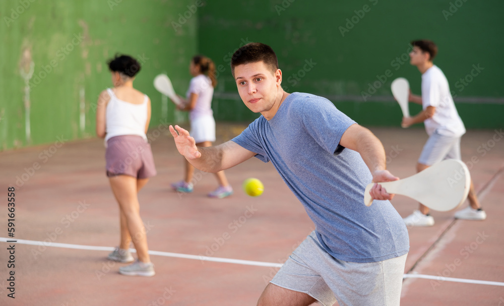 Focused young man playing friendly pelota goma match on outdoor summer court with fronton wall, swinging wooden paleta to hit small rubber ball
