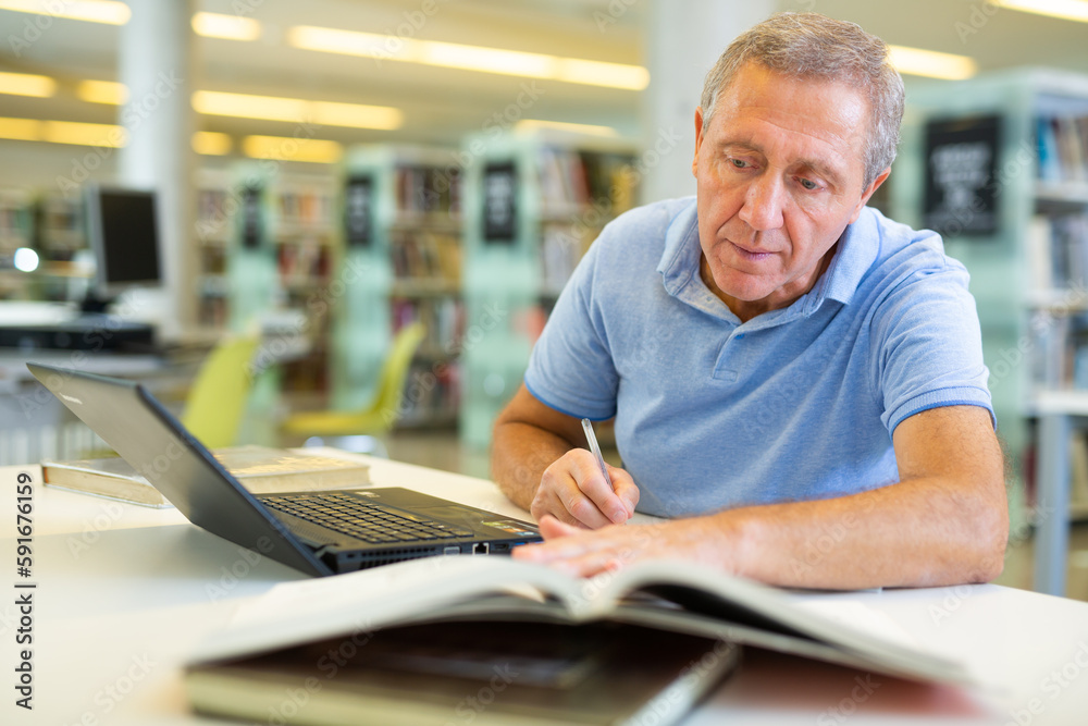 Fototapeta premium Concentrated mature man spending time in library, reading books and making laptop. Self-education concept
