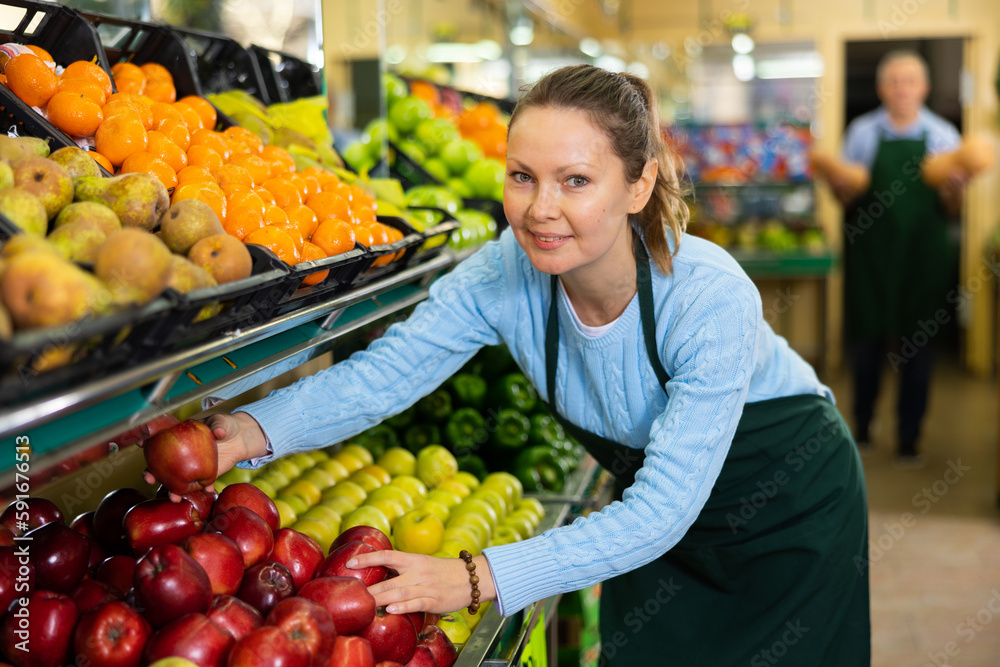 Friendly middle-aged female seller in apron arranging red delicious apples during workday in supermarket