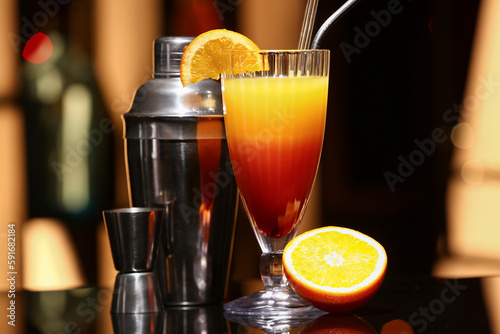 Glass of tasty Tequila Sunrise, cocktail shaker and double jigger on dark table in bar