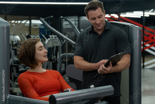 Young Caucasian woman doing workout in the gym for her strength with the advisory from her trainer.She is wearing red exercise top outfit and and hands on the workout machine.
