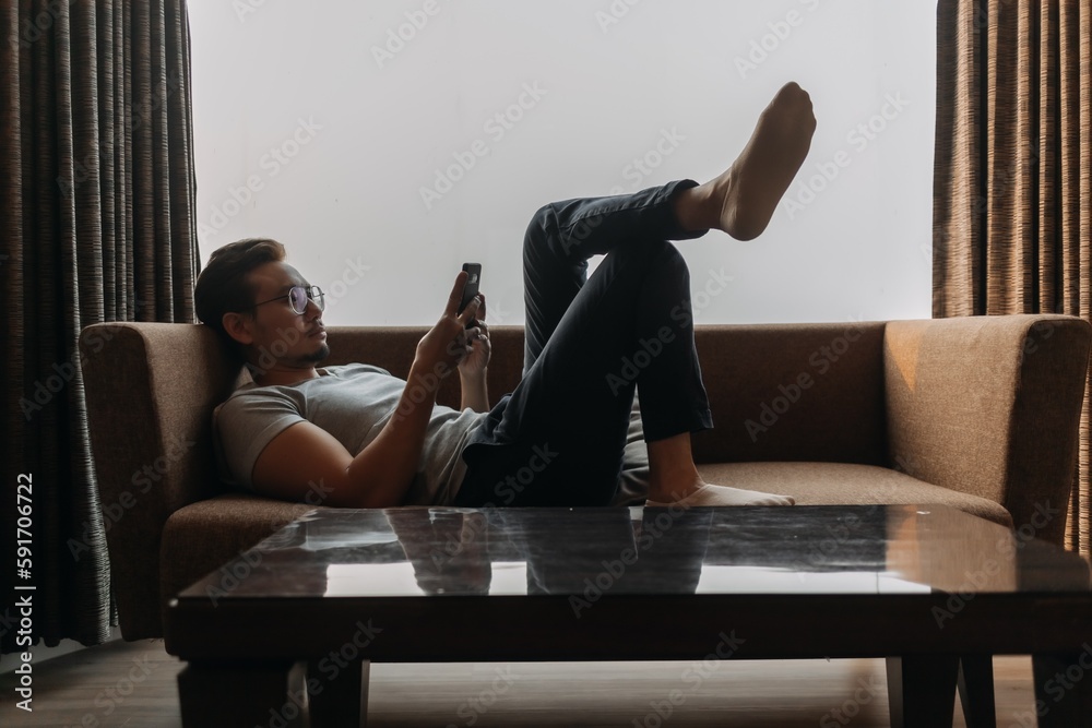 Asian man lies on sofa using phone with his feet on the table. Concept of manner Asian man lies on sofa using phone with his feet on the table. Concept of manner