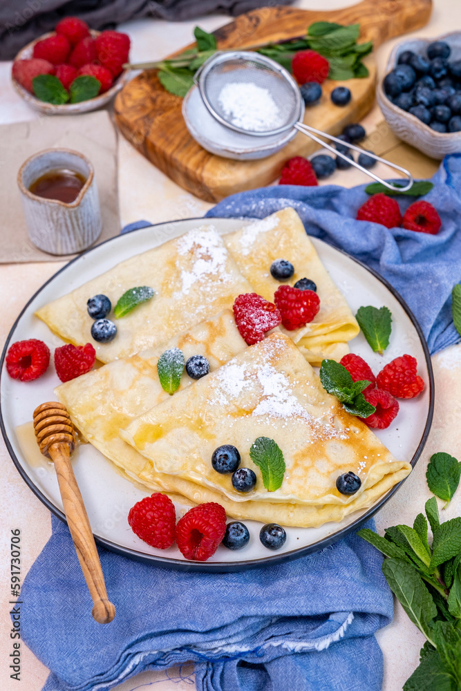 Crepes garnished with blueberries, raspberries, fresh mint leaves and powdered sugar served on a round plate. Crepes garnished with blueberries, raspberries, fresh mint leaves and powdered sugar served on a round plate.