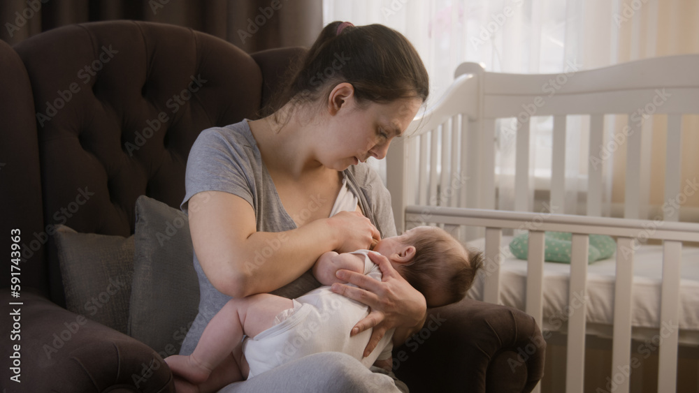 Caring mother breastfeeds little newborn child, sits on armchair near crib. Woman holds baby daughter or son in her arms while feeding with breast milk. Concept of childhood, motherhood and family. Caring mother breastfeeds little newborn child, sits on armchair near crib. Woman holds baby daughter or son in her arms while feeding with breast milk. Concept of childhood, motherhood and family.