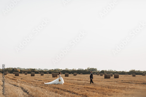 Wedding portrait of the bride and groom. Bride and groom on the background of hay, walk across the field to meet each other. Red-haired bride in a long dress. Stylish groom. Summer. Bales of hay