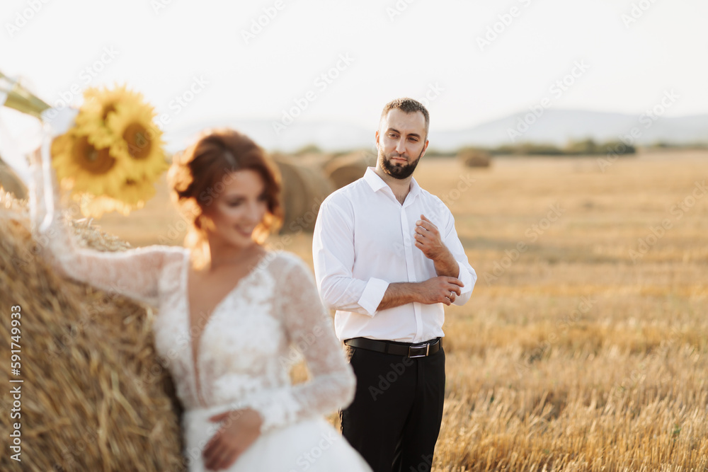 Wedding portrait of the bride and groom. The groom, tearing his shirt, stands behind the bride, near a bale of hay. Red-haired bride in a long dress with a bouquet of sunflowers. Stylish groom. Summer Wedding portrait of the bride and groom. The groom, tearing his shirt, stands behind the bride, near a bale of hay. Red-haired bride in a long dress with a bouquet of sunflowers. Stylish groom. Summer