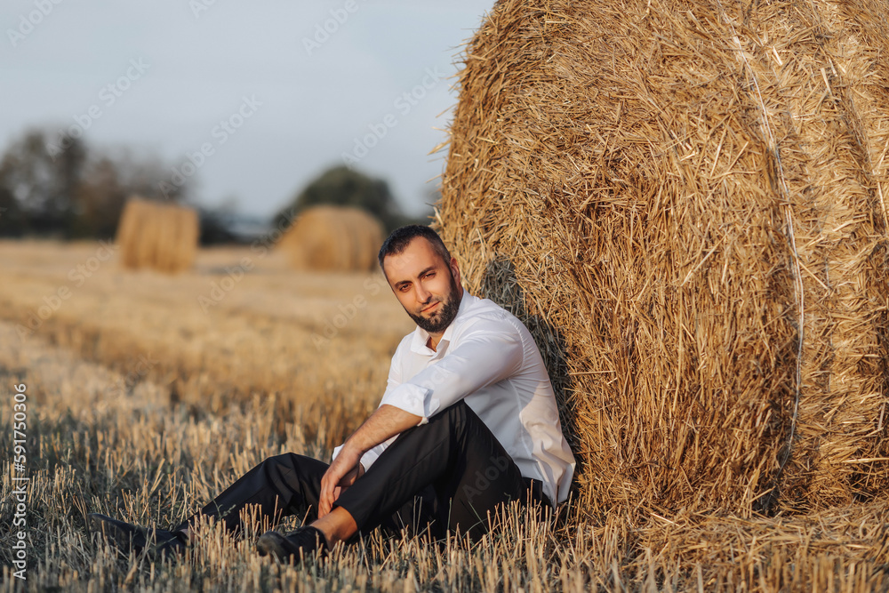 Wedding portrait photo. A stylish groom in a white shirt sits relaxed near a hay bale and looks into the lens. Bearded man. Style. Wedding portrait photo. A stylish groom in a white shirt sits relaxed near a hay bale and looks into the lens. Bearded man. Style.