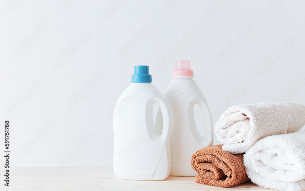 Washing gel liquid laundry detergent and fabric softener, pile of towels on a wooden table against a light white background with copy space.