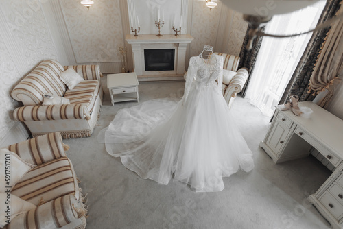 an exquisite wedding dress with a long train on a mannequin is located in a hotel room with a royal interior. Wide angle © Vasil