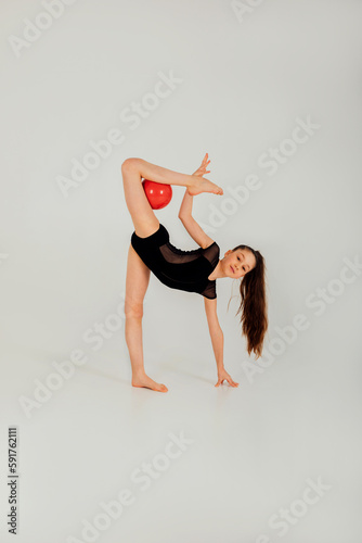 Artistic gymnast girl in a black leotard with a red ball does tricks on a white background. Front view