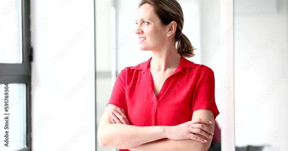 Positive female manager in red blouse turns to look out bright window. Happy woman stands in office with crossed arms smiling slow motion Positive female manager in red blouse turns to look out bright window. Happy woman stands in office with crossed arms smiling slow motion