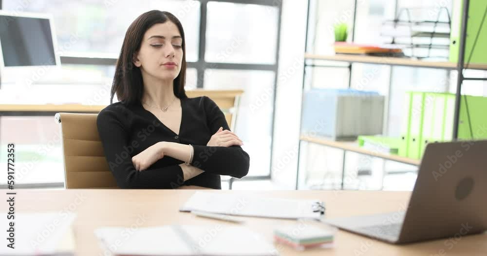 Tired secretary crosses arms leaning on back of chair at workplace. Young brunette woman sits at table near open laptop in office slow motion Tired secretary crosses arms leaning on back of chair at workplace. Young brunette woman sits at table near open laptop in office slow motion