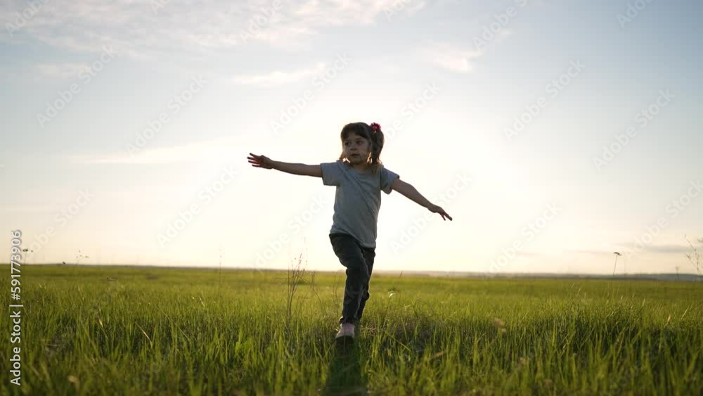 Happy child run her arms to sides. Girl play in park at sunset. Little girl run through green grass in the field. Dream girl. Cheerful child runs smiling and dreaming. Winner girl strives for success Happy child run her arms to sides. Girl play in park at sunset. Little girl run through green grass in the field. Dream girl. Cheerful child runs smiling and dreaming. Winner girl strives for success
