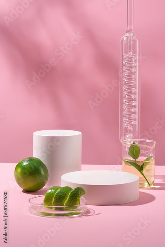 Background for the presentation of cosmetic products with white cylinder podium  lime  peel in petri dish and liquid in beaker  on pink background with natural shadow leaves. Front view  copy space.