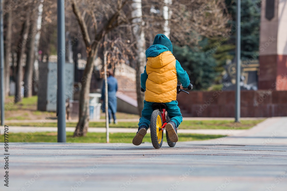 Obraz premium A cheerful little boy rides a bicycle outdoors. A happy child walks in the spring park. The baby is dressed in a fashionable yellow vest and turquoise jumpsuit.