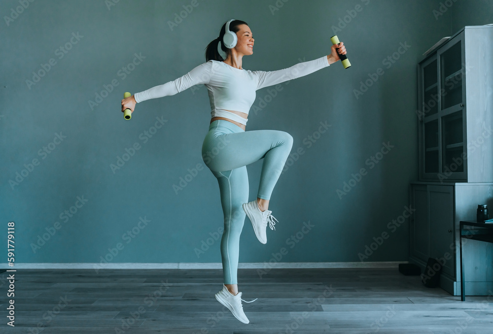 Active brunette American girl in sportswear jumps at home holds dumbbells, at morning workout indoors enjoys music uses headphones. Gym, fitness healthcare. Pretty fit young woman trains at sport club