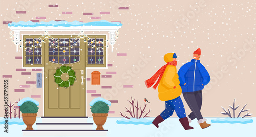 People wearing warm winter clothes walk down city street outside in cold season. Woman and man walking in snowy weather at house wall with entrance door decorated with Christmas wreath and decorations