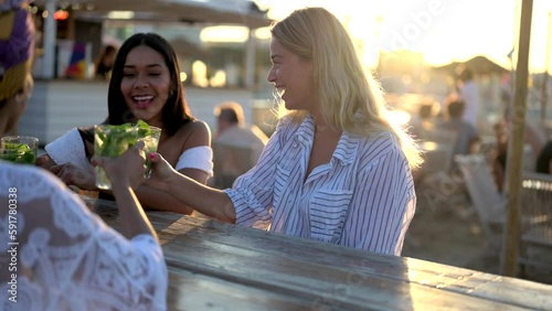 Young beautiful women drinking and chatting at cocktail party on the beach, female friends having fun at chiringuito during vacation, holiday activities, slow motion clip photo