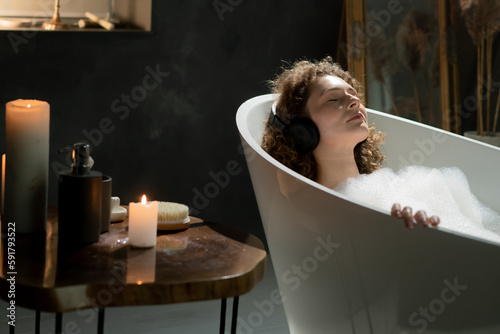 Photo of a beautiful woman who lies in the bath with her eyes closed and listens to music through wireless headphones. A young woman relaxes in a bath with aromatic candles and listens to music on