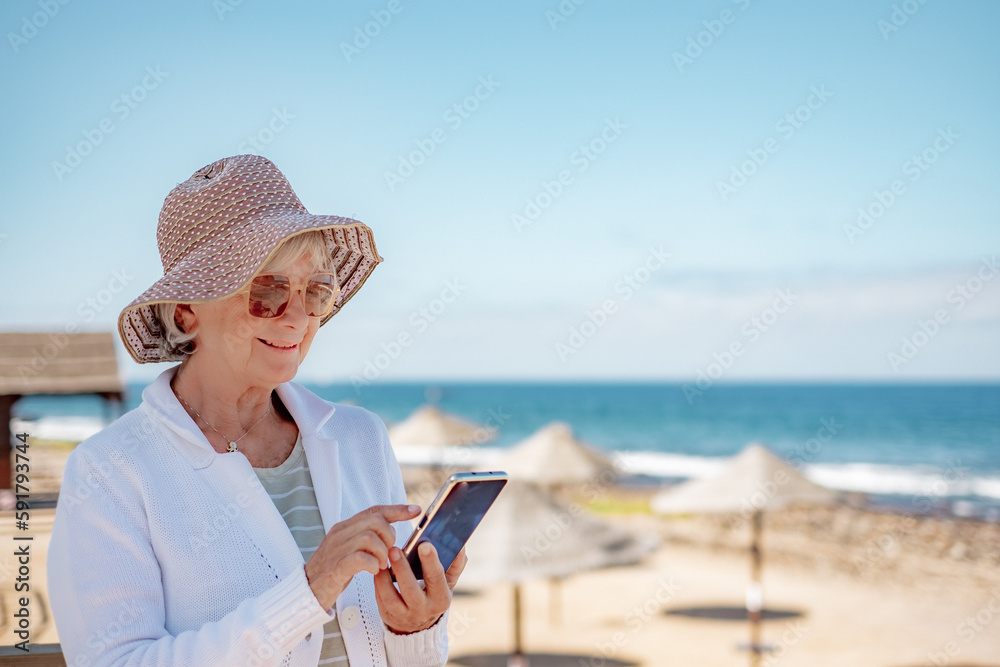 Smiling senior woman wearing hat and eyeglasses standing at the beach using mobile phone typing message, horizon over sea and blue sky