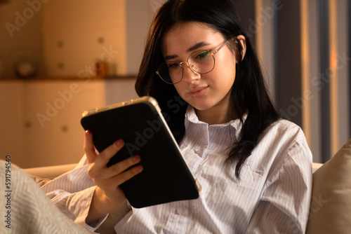 Cute woman in glasses uses a tablet while lying on the sofa. The long-haired brunette spends her evening watching TV shows on her digital tablet.