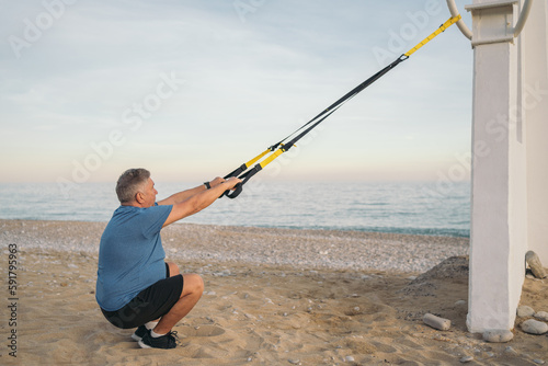 Side image of an older, white-haired, overweight gentleman squatting squatting down while gripping the straps of a suspension training system on the beach.